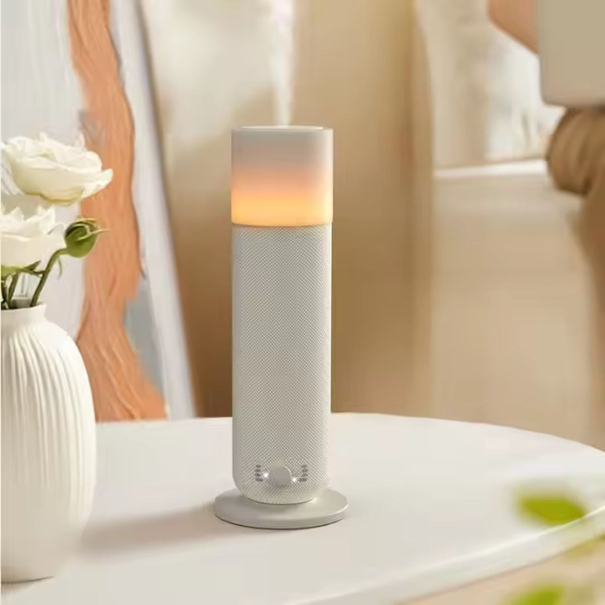 A white cylindrical diffuser or humidifier with a glowing warm light on top, sitting on a white table next to a vase of white roses in a softly lit indoor setting.
